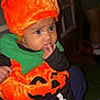 baby, child, pumpkin_costume, orange_hat, green_collar, cute, indoors, person, face, costume, holiday, halloween, plush, black_clothing, expression, finger_in_mouth, wide_eyes, sitting, legs, adult_hand