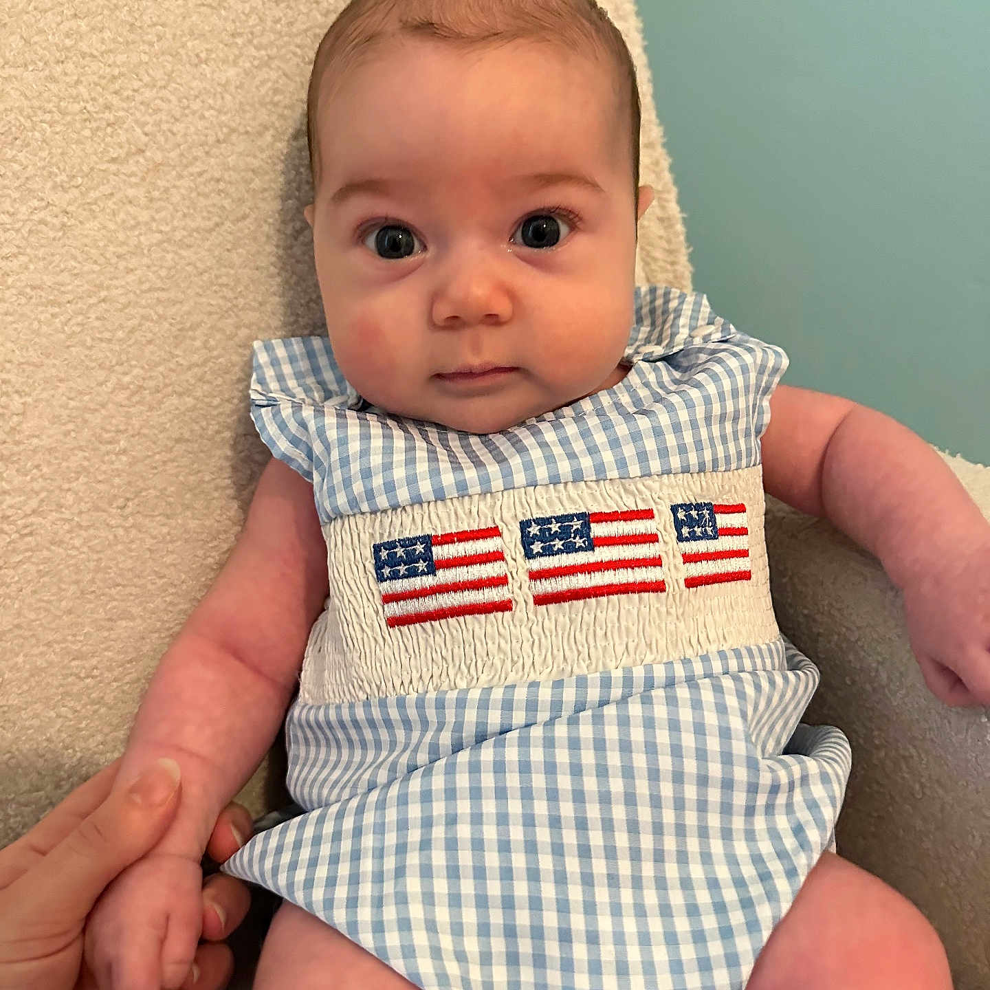 Elijah is registered to the contest to win money with this photo: american_flag, baby, background, beige, blue_gingham, chair, child, cute, embroidery, eyes, face, fingers, hand_holding, indoor, infant, person, portrait, romper, skin, texture