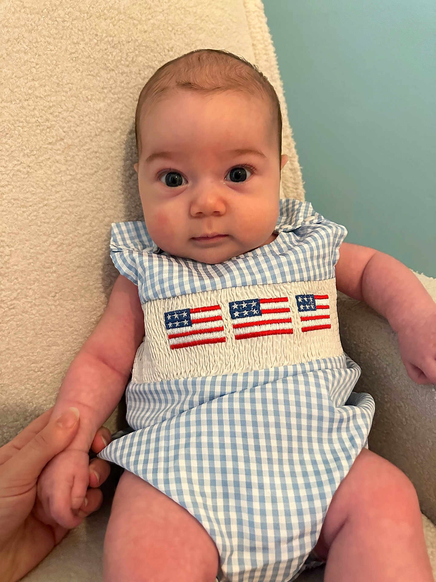Elijah is registered to the contest to win money with this photo: baby, child, infant, blue_gingham, romper, american_flag, embroidery, hand_holding, chair, beige, texture, person, portrait, cute, indoor, skin, face, eyes, fingers, background
