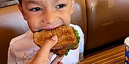 Noah is registered to the contest to win money with this photo: child, boy, sandwich, hand, restaurant, booth_seating, table, fries, cup, food, casual_dining, eating, smiling, indoor, clothing, white_shirt, lettuce, bread, person, happy