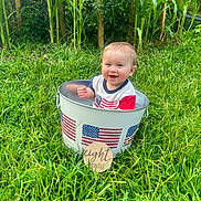 Ian is registered to the contest to win money with this photo: baby, bucket, grass, outdoor, greenery, corn_stalks, smiling, child, happy, american_flag, sign, months, nature, summer, portrait, infant, clothing, metal_bucket, sunlight, playful
