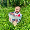 baby, bucket, grass, outdoor, greenery, corn_stalks, smiling, child, happy, american_flag, sign, months, nature, summer, portrait, infant, clothing, metal_bucket, sunlight, playful