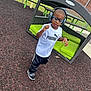 bench, black_pants, boy, brick_wall, casual, child, fence, glasses, grass, green_cushion, headphones, mulch, music, outdoor, picnic_table, playground, puma, smiling, sneakers, white_shirt