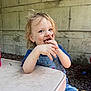 blue_clothing, child, concrete_wall, face, food, ground, hair, hands, happy, light, messy, outdoor, overalls, seat, shadow, smiling, snack, table, toddler, umbrella