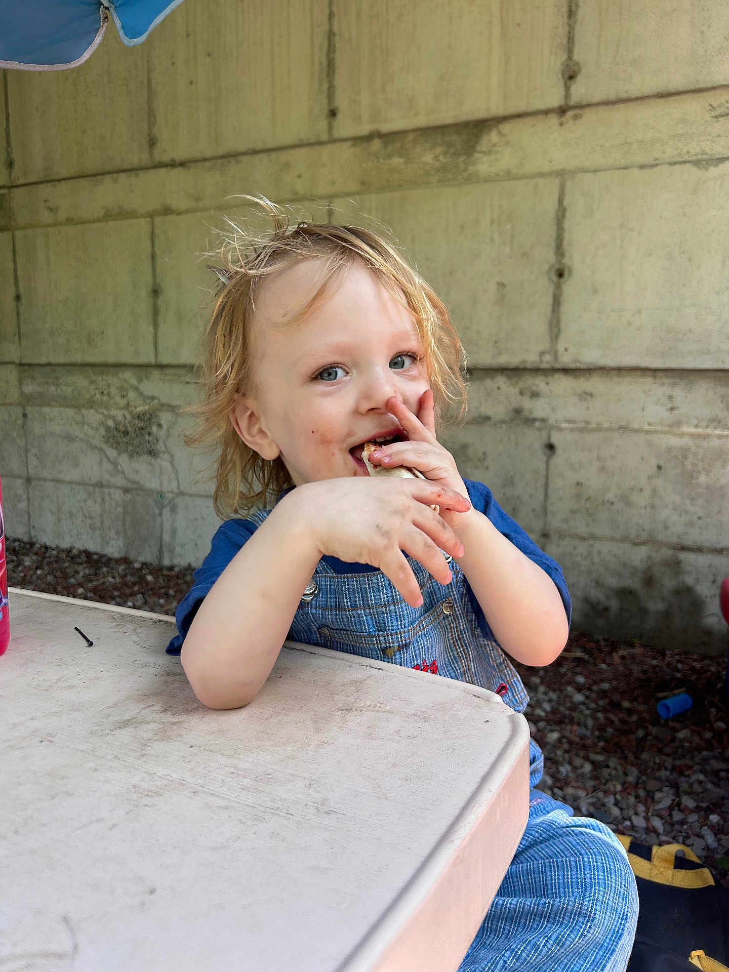 Paxton joined the competition — help win amazing prizes! blue_clothing, child, concrete_wall, face, food, ground, hair, hands, happy, light, messy, outdoor, overalls, seat, shadow, smiling, snack, table, toddler, umbrella