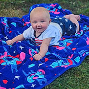 Eros is registered to the contest to win money with this photo: baby, child, blanket, blue_blanket, cartoon_characters, grass, outdoor, smiling, happy, toddler, cute, person, clothing, shorts, shirt, nature, lying_down, playful, skin, head