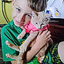 boy, child, cat, pet, indoor, green_shirt, pink_clothing, smiling, holding, furniture, television, background, metal_headboard, short_hair, cute, animal, person, happy, home, casual