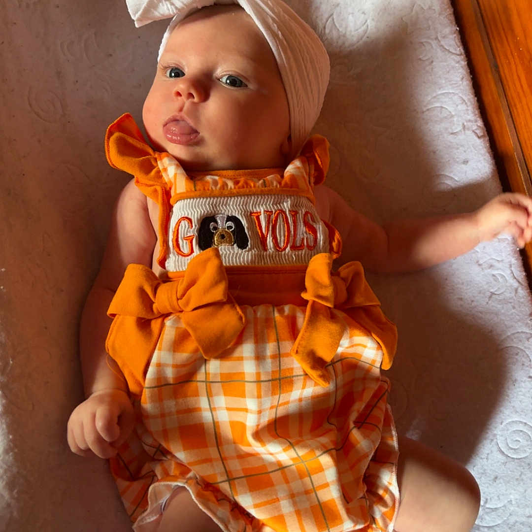 Eliana is registered to the contest to win money with this photo: baby, child, headband, bow, orange_clothing, romper, plaid, blanket, floor, wooden_floor, indoor, cute, infant, person, lying_down, expression, tongue_out, texture, soft, casual