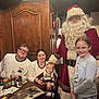 family, indoor, santa_claus, child, baby, woman, man, glasses, pajamas, table, food, book, holiday, christmas_decorations, smile, beard, pacifier, wooden_paneling, festive, celebration