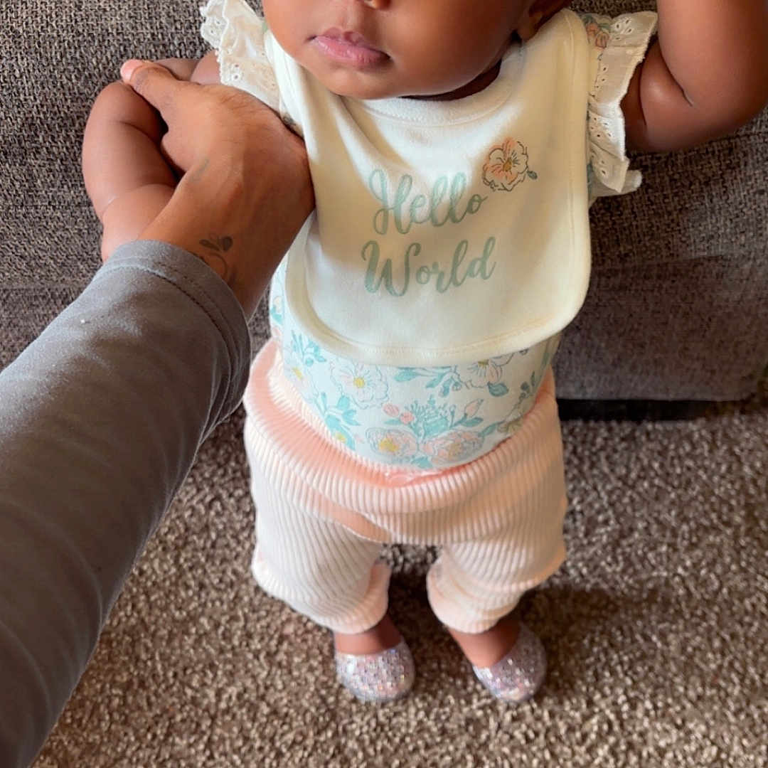 Omiyah is registered to the contest to win money with this photo: baby, child, infant, person, face, hand, clothing, bib, headband, carpet, floor, pacifier, toy, standing, support, cute, expression, indoors, home, footwear