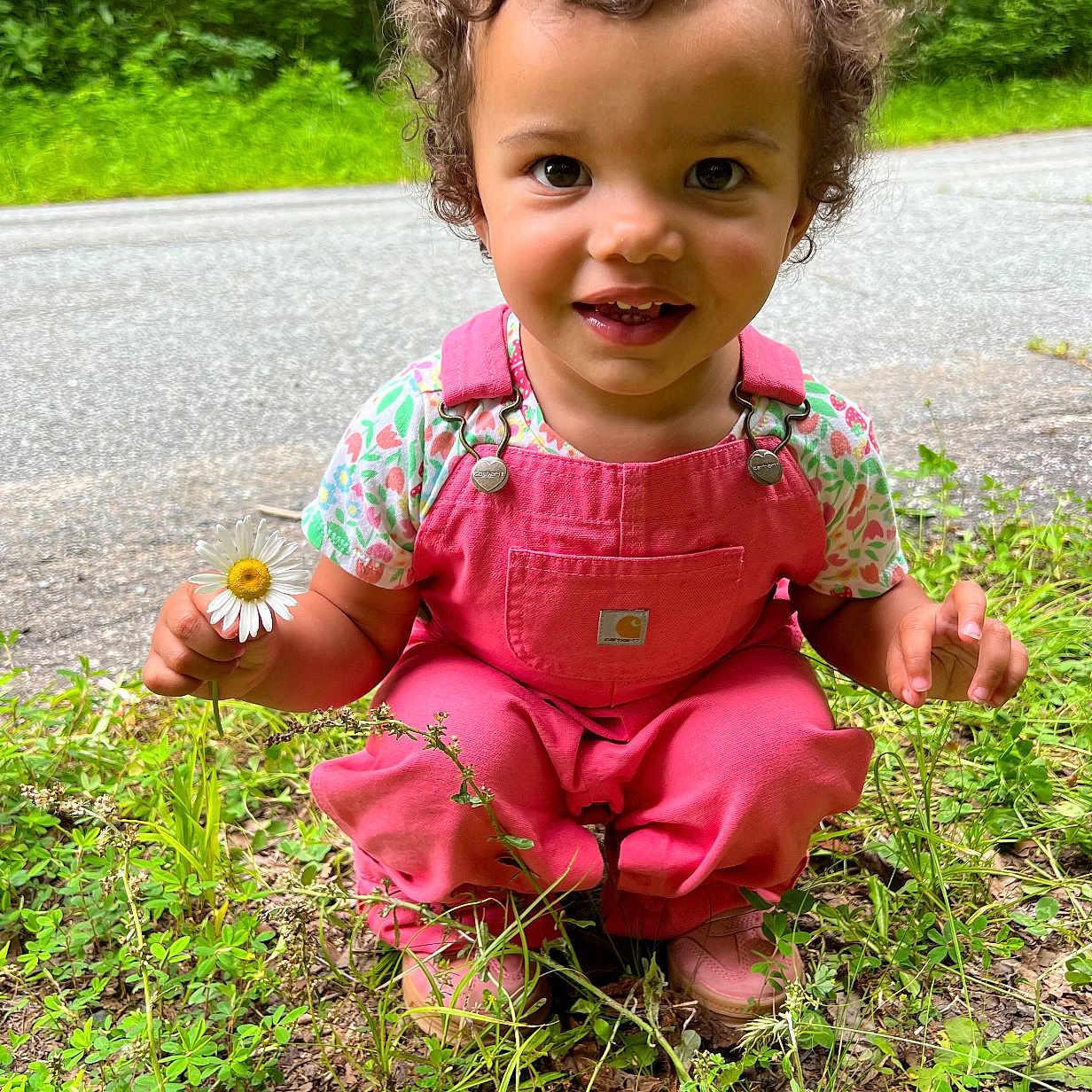 Deava joined the competition — help win amazing prizes! child, clothing, curly_hair, cute, daisy, daylight, eyes, face, flower, grass, greenery, happy, nature, outdoor, person, pink_overalls, portrait, road, smiling, toddler