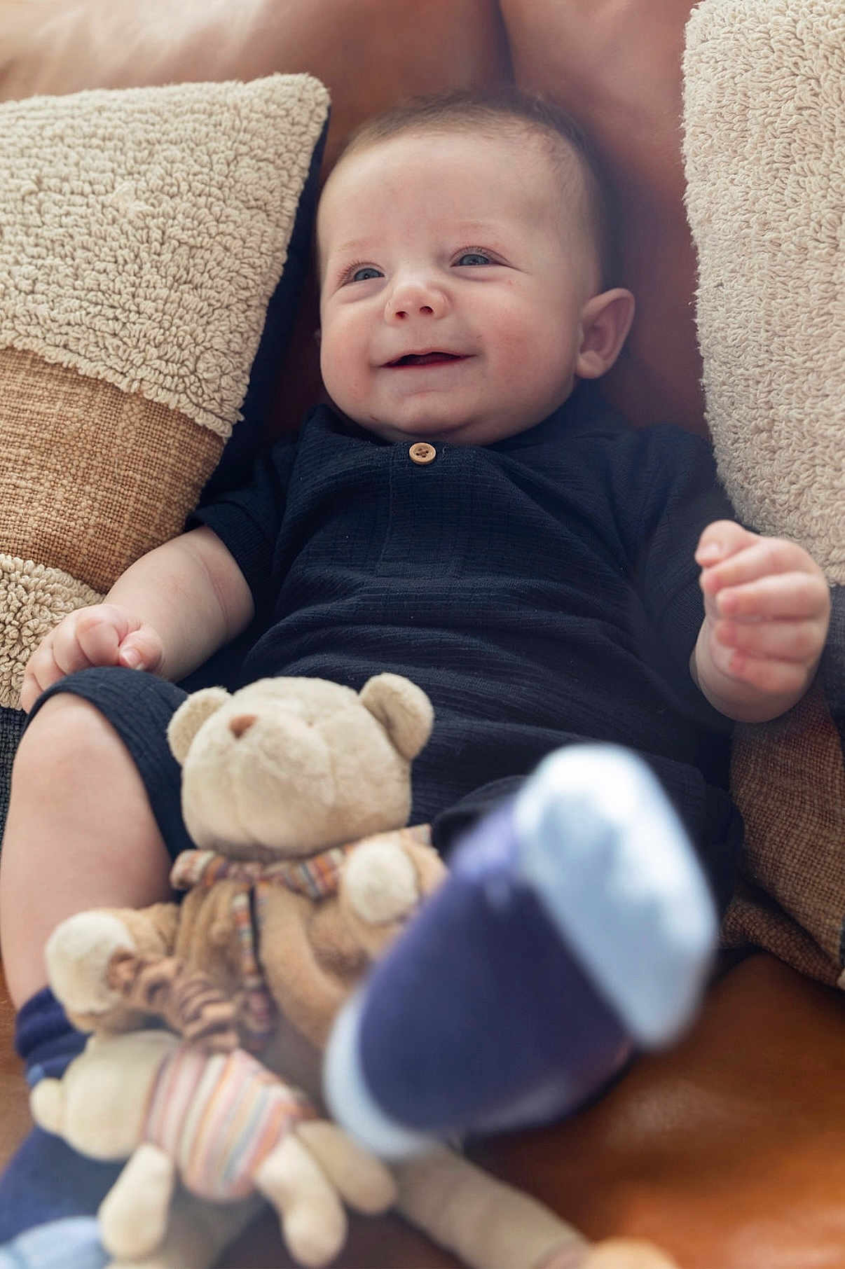 Leandros is registered to the contest to win money with this photo: baby, infant, smile, face, pillow, couch, plush_toy, teddy_bear, clothing, footwear, happy, child, sitting, indoor, furniture, soft_toy, cute, comfort, portrait, relaxation