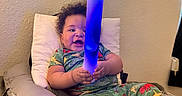 Oko is registered to the contest to win money with this photo: baby, child, pajamas, dinosaur_print, glow_stick, light, colorful, cushion, pillow, indoor, smiling, happy, sitting, curly_hair, foot, hand, wall, bed, room, person