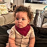 baby, child, curly_hair, indoor, blanket, couch, bib, clothing, furniture, television, living_room, seated, portrait, cute, infant, expression, soft_lighting, home, person, cozy