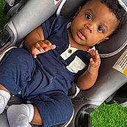 Noa is registered to the contest to win money with this photo: baby, child, infant, car_seat, blue_outfit, white_socks, curly_hair, brown_skin, indoors, seat_belt, gray_fabric, green_grass, cute, portrait, looking_up, hands, expression, comfortable, casual_clothing, young