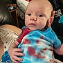 baby, child, person, tie_dye, shirt, hand, face, blue, red, indoor, couch, pillow, clothing, pacifier, skin, expression, looking, holding, furniture, home