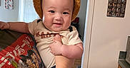 Zaire is registered to the contest to win money with this photo: baby, child, infant, portrait, smiling, happy, bear_hat, orange_hat, costume, held, indoors, home, ceiling_fan, window, doorway, wall_decor, cocktail_chart, cartoon_shirt, adult_arm, family