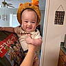 baby, child, infant, portrait, smiling, happy, bear_hat, orange_hat, costume, held, indoors, home, ceiling_fan, window, doorway, wall_decor, cocktail_chart, cartoon_shirt, adult_arm, family