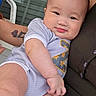 Zaire is registered to the contest to win money with this photo: baby, child, infant, smiling, portrait, sitting, onesie, gray_clothing, outdoor, patio, chair, couch, adult, lap, face, eyes, mouth, tattoo, cooler, photography
