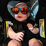 Zaire is registered to the contest to win money with this photo: baby, child, infant, car_seat, sunglasses, smiling, sitting, buckled_in,  안전, black_clothing, portrait, close_up, vehicle, harness, indoor, face, hands, legs, reflective_lenses, cute