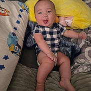 Zaire is registered to the contest to win money with this photo: baby, child, infant, smiling, tongue_out, portrait, sitting, lying_down, bed, blanket, pillow, yellow_pillow, plush_toy, checkered_clothing, onesie, cartoon_print, indoor, bedroom, happy, photography