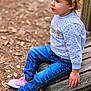 casual_clothing, child, curly_hair, daylight, ground, jeans, leaves, nature, outdoor, pine_needles, pink_sneakers, playground, portrait, side_view, sitting, sweater, thoughtful, toddler, wooden_bench, young_child