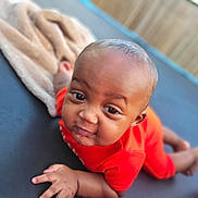 Kaizen is registered to the contest to win money with this photo: baby, infant, orange_clothing, trampoline, blanket, outdoor, fence, face, head, hand, skin, person, child, playful, expression, lying_down, closeup, daylight, smile, curious