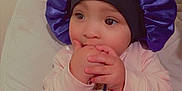 Juniper is registered to the contest to win money with this photo: baby, child, clothing, cozy, curious, cute, expression, face, hand_in_mouth, headwear, indoor, infant, person, pink_outfit, portrait, purple_bonnet, sitting, soft_lighting, toddler, young_child