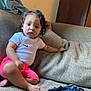 toddler, child, curly_hair, white_tshirt, pink_shorts, sofa, couch, indoor, living_room, furniture, barefoot, person, young_child, casual_clothing, relaxed, seated, home, comfort, expression, portrait