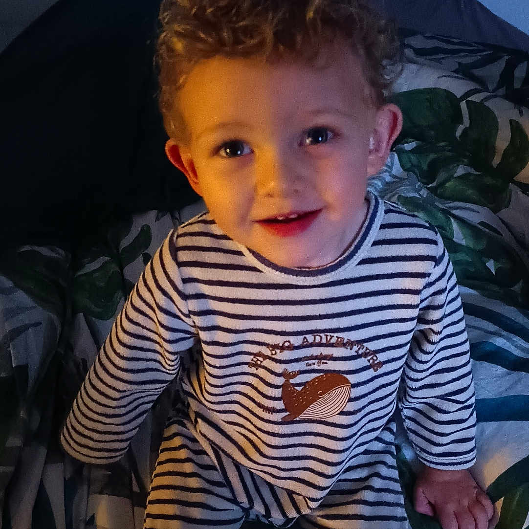 Aryson participe au concours pour gagner de l'argent avec cette photo : bed, bedspread, child, cozy, curly_hair, cute, evening_light, face, happy, home, indoor, leaf_pattern, pajamas, person, portrait, smiling, striped_pajamas, toddler, whale_print, young_child