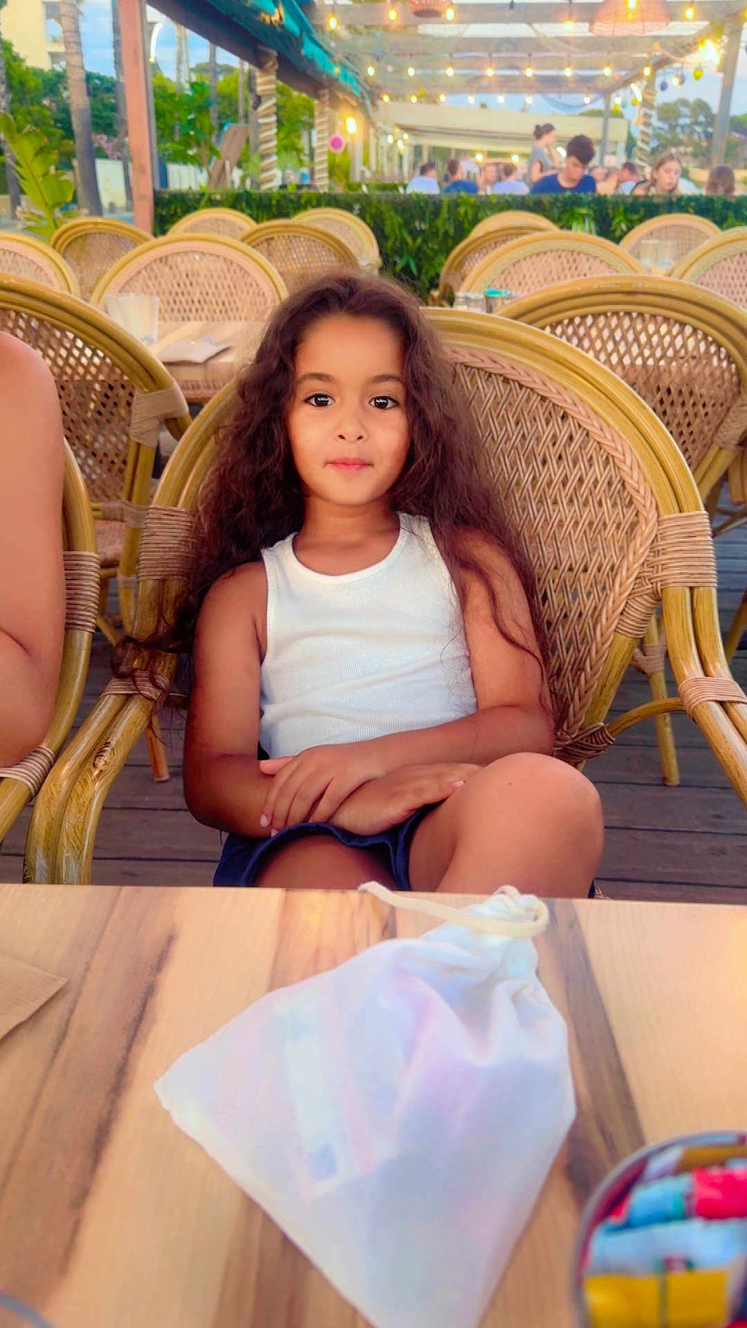 Ritej a rejoint le concours — aidez-le/la à gagner de superbes lots ! child, girl, long_hair, curly_hair, white_tank_top, blue_skirt, wicker_chair, outdoor_cafe, wooden_table, drawstring_bag, string_lights, evening, people_in_background, relaxed, summer, casual_clothing, seated, portrait, daylight, restaurant