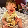 Emberly is registered to the contest to win money with this photo: toddler, child, smiling, burger, bed, yellow_onesie, curly_hair, indoor, clothing, happy, person, furniture, blanket, foot, toes, playpen, pile_of_clothes, face, expression, sitting