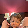 toddler, child, phone, purple_shirt, floral_pattern, bedroom, lamp, red_wall, headboard, curly_hair, person, indoor, legs, tattoo, expression, holding, communication, closeup, cute, baby