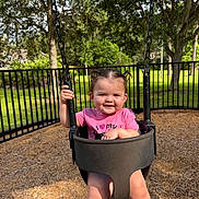 Georgia is registered to the contest to win money with this photo: toddler, child, swing, playground, outdoor, smile, happy, grass, tree, fence, sunlight, pigtails, pink_shirt, park, wood_chips, person, baby, nature, daytime, fun