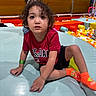 child, toddler, curly_hair, red_shirt, jordan, orange_socks, ball_pit, play_area, indoor, padded_surface, colorful, casual_clothing, young_child, curious_expression, fun, kid, portrait, leisure, bright_colors, active