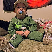 Noah is registered to the contest to win money with this photo: toddler, child, pacifier, cap, green_clothing, dinosaur_print, indoor, carpet, blanket, tapestry, shoe, sitting, cute, face, person, footwear, casual, young_child, home, relaxed