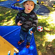 Noah joined the competition — help win amazing prizes! blue_mat, boy, child, cute, daytime, dinosaur_sweater, grass, happy, hat, jeans, nature, outdoor, pavement, playground, playing, shoes, soccer_ball, sunlight, toy_trucks, young_child