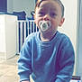 toddler, child, pacifier, blue_sweater, blue_pants, indoor, kitchen, wooden_toy, walker, baby_gate, floor_tiles, cabinet, child_hands, child_face, person, home, baby, young_child, furniture, household