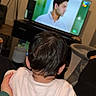child, toddler, baby, back_view, sitting, television, watching_tv, living_room, indoor, home, adult_hand, support, short_hair, pink_shirt, furniture, floor_lamp, wall_clock, cozy, entertainment, family