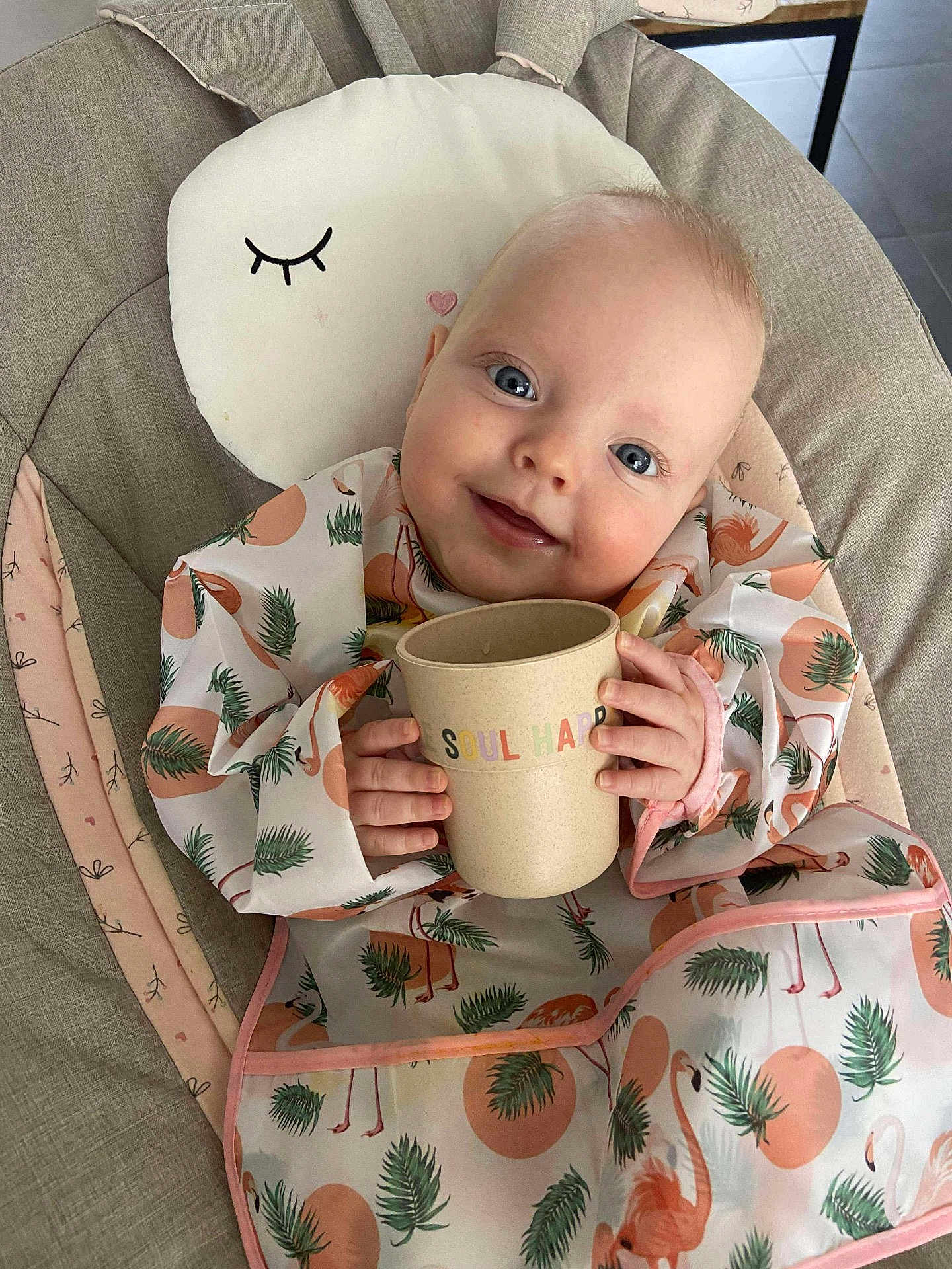 Emmie participe au concours pour gagner de l'argent avec cette photo : baby, child, smiling, bib, cup, flamingo_pattern, cushion, chair, indoor, pillow, cute, happy, person, infant, clothing, face, hand, head, blue_eyes, blanket