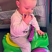 Brandon is registered to the contest to win money with this photo: baby, child, toy_car, plastic_toy, indoor, carpet, playing, chewing, purple, green, yellow, sitting, cute, infant, clothing, long_sleeve, pants, face, hand, foot