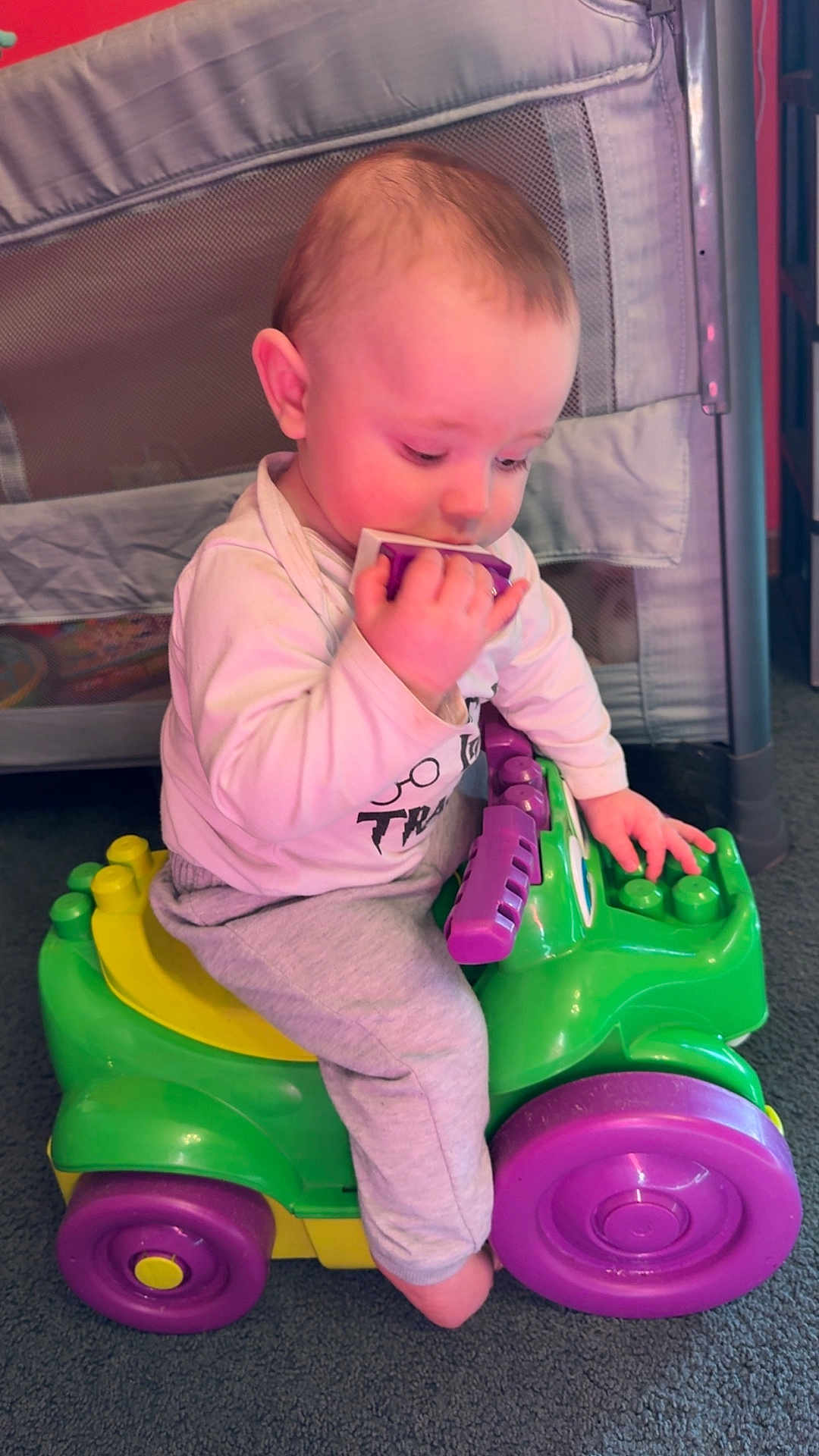 Brandon is registered to the contest to win money with this photo: baby, child, toy_car, plastic_toy, indoor, carpet, playing, chewing, purple, green, yellow, sitting, cute, infant, clothing, long_sleeve, pants, face, hand, foot