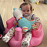 baby, blue_eyes, bright, colorful, cute, holding, infant, inflatable_seat, onesie, pacifier, patterned_pajamas, pink, playroom, plush_toy, ponytail, portrait, sitting, soft_texture, toy_bin, wooden_floor