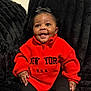 baby, child, smiling, red_fleece, black_bow, sitting, couch, indoor, happy, portrait, person, furniture, black_pants, cute, young_child, warm_clothing, cozy, bright_color, expression, infant