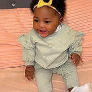 Kemora is registered to the contest to win money with this photo: baby, child, infant, smiling, sitting, headband, yellow_bow, curly_hair, mint_green_clothing, long_sleeves, patterned_outfit, bed, pink_bedspread, happy, indoors, cute, sock, soft_texture, person, portrait