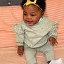 baby, child, infant, smiling, sitting, headband, yellow_bow, curly_hair, mint_green_clothing, long_sleeves, patterned_outfit, bed, pink_bedspread, happy, indoors, cute, sock, soft_texture, person, portrait