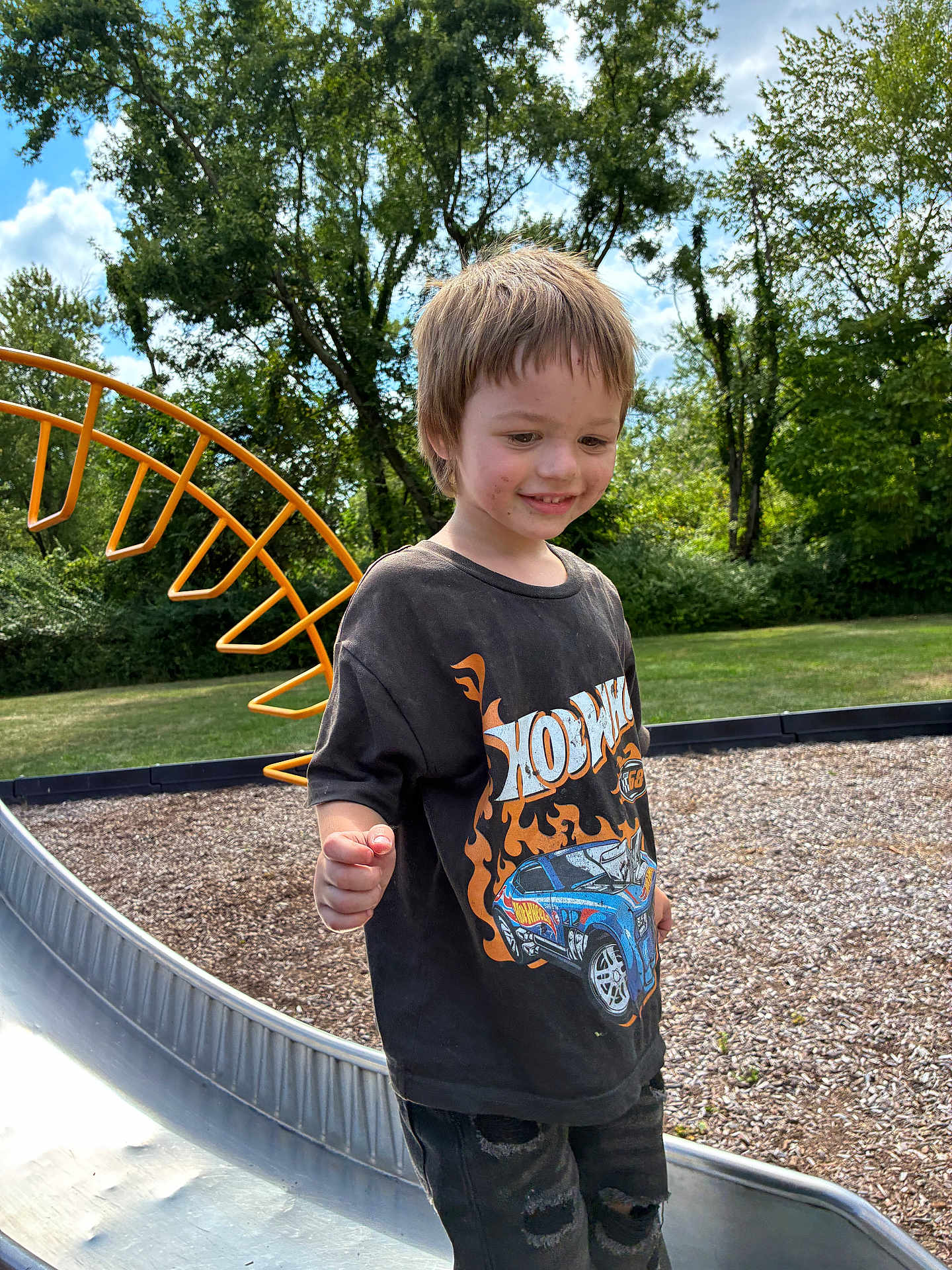 Uhtred is registered to the contest to win money with this photo: child, boy, playground, slide, hot_wheels, tshirt, ripped_jeans, outdoor, trees, grass, sky, clouds, smiling, dirt, summer, fun, leisure, casual_clothing, daytime, happy