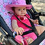 baby, cat_pattern, child, cute, expression, fabric, footwear, hand, infant, nature, outdoor, pink_clothing, pink_hat, playful, safety_harness, seat, stroller, summer, sunlight, toy
