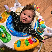 Anniliah joined the competition — help win amazing prizes! baby, child, infant, smiling, happy, toy, activity_center, playtime, colorful, floor, wooden_floor, clothing, black_outfit, skeleton_pattern, face, person, indoor, cute, young, curious