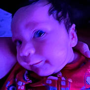Adrin joined the competition — help win amazing prizes! baby, infant, face, closeup, child, red_clothing, fire_truck_print, curly_hair, indoors, portrait, young_child, cute, soft_lighting, unfocused, blurry, head, person, expression, purple_tint, hand